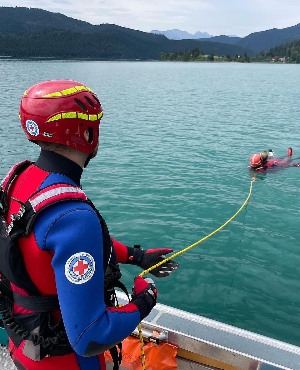 Wasserrettung vom Boot aus. Ein Wasserwachtler hält eine schnur, an der sich ein weiterer Wasserwachtler und ein hilfloser Schwimmer geborgen wird.